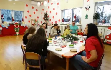 Na krzesełkach przy stolikach siedzą dorośli z dziećmi na kolanach. Tuż przed nimi leżą drewniane krążki i materiały do dekoracji świątecznych.