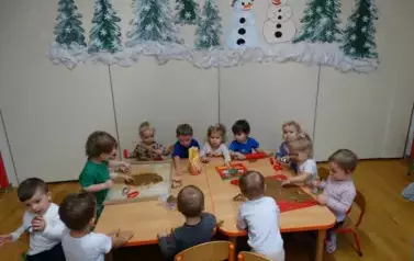 Przy stoliku siedzą dzieci. Tuż przed nimi leżą dwie stolnice z brązową masą. W tle zielone choinki i białe bałwanki.