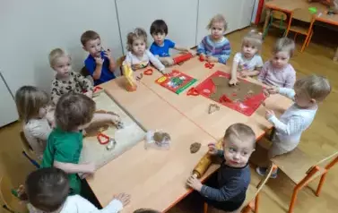 Przy stoliku siedzą dzieci, w ręce trzymają foremki do wycinania. Tuż przed nimi leżą dwie stolnice z brązową masą. 