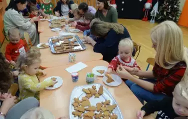 Grupa dorosłych z dziećmi siedzi przy stolikach. Tuż przed nimi leżą białe talerzyki a na nich pierniczki w kształcie choinek, gwiazdek, bałwank&oacute;w. W tle sala zabaw z bożonarodzeniową dekoracją. 