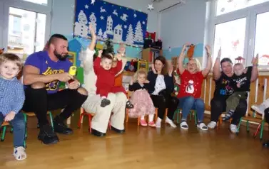 Sala zabaw-na krzesełkach siedzą dorośli z dziecmi na kolanach. Wszyscy mają uniesione ręce ku g&oacute;rze.