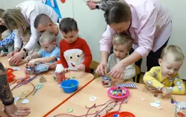Przy stoliku siedzi chłopiec. obok stoi osoba dorosła. Ich ręce dotykają słoiczka stojącego na stole. W tle grupa dorosłych z dziećmi.