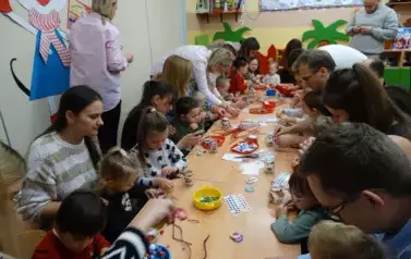 Grupa dorosłych os&oacute;b z dziećmi siedzi przy stolikach, na kt&oacute;rych leżą materiały do świąteczne dekoracji.