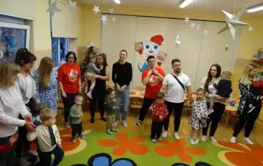 Grupa dorosłych os&oacute;b z dziećmi stoi na dywanie, w tle srebrzyste gwiazdki.