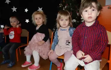 Na tle ciemnej ściany z błyszczącymi gwiazdkami siedzi grupka dzieci w kolorowych ubrankach.