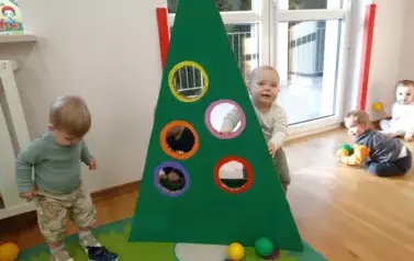 Na podłodze stoi tr&oacute;jkątna zielona choinka z okrągłymi otworami. Dziecko dotyka ręką otworu w choince.