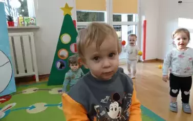 Na pierwszym planie buzia dziecka, w tle inne dzieci stojące przy choince.