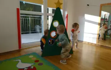Na podłodze stoi tr&oacute;jkątna zielona choinka z okrągłymi otworami. Dziecko trzyma w ręku małą piłeczkę i dotyka otworu w choince. 