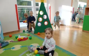 Na podłodze stoi tr&oacute;jkątna zielona choinka z okrągłymi otworami. Wok&oacute;ł niej stoją dzieci.