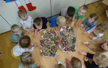 Przy stoliku stoją dzieci. Tuż przed nimi leżą dwie tace z kolorowymi piernikami.