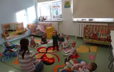 Sala zabaw-dzieci z osobą dorosłą siedzą na kolorowym dywanie.