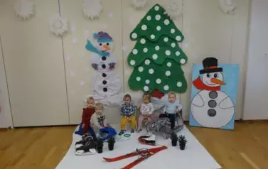Sala zabaw-grupka dzieci siedzi na sankach. W tle zielona choinka i dwa bałwanki.