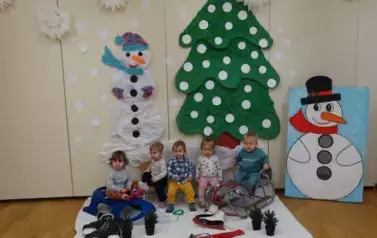 Sala zabaw-grupka dzieci siedzi na sankach. W tle zielona choinka i dwa bałwanki.