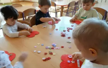 Sala zabaw-grupka dzieci siedzi przy stoliku. Tuż przed nimi leżą czerwone serduszka i kolorowe kwiatuszki.
