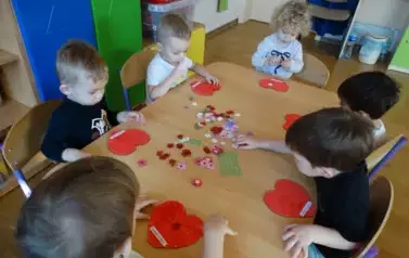 Sala zabaw-grupka dzieci siedzi przy stoliku. Tuż przed nimi leżą czerwone serduszka i kolorowe kwiatuszki.