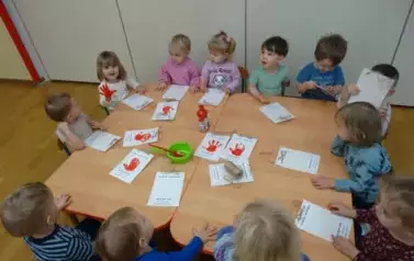 Sala zabaw-grupka dzieci siedzi przy stoliku. Jedna dziewczynka siedzi z uniesioną czerwoną dłonią. Tuż przed nimi leżą białe kartki papieru z odbitą czerwoną rączką.