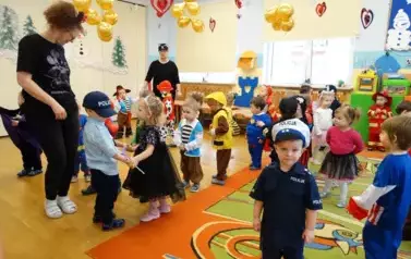 Sala zabaw-grupa dzieci i Opiekunowie w balowych strojach stoją na podłodze. W tle złote balony i czerwone serduszka.