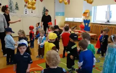 Sala zabaw-grupa dzieci i Opiekunowie w balowych strojach stoją na podłodze. W tle złote balony i czerwone serduszka.