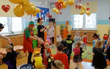 Sala zabaw-grupa dzieci i Opiekunowie w balowych strojach stoją na podłodze. W tle kolorowe balony i czerwone serduszka.