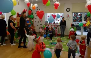 Sala zabaw-grupa dzieci i Opiekunowie w balowych strojach stoją na dywanie. W tle kolorowe balony i czerwone serduszka.