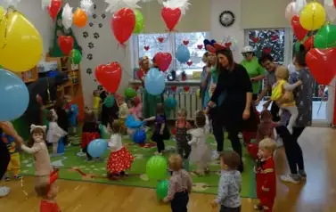 Sala zabaw-grupa dzieci i Opiekunowie w balowych strojach stoją na dywanie. W tle kolorowe balony, białe gwiazdy i czerwone serduszka.