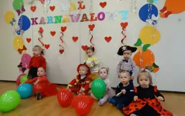 Sala zabaw-na tle jasnej ściany siedzi grupka dzieci w balowych strojach, a na niej kolorowe balony, czerwone serduszka i napis BAL KARNAWAŁOWY.