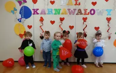 Sala zabaw-na tle jasnej ściany stoi grupka dzieci w balowych strojach, a na niej kolorowe balony, czerwone serduszka i napis BAL KARNAWAŁOWY.