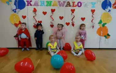 Sala zabaw-grupka dzieci w balowych strojach stoi na dywanie. W tle kolorowe balony, czerwone serduszka i napis BAL KARNAWAŁOWY.