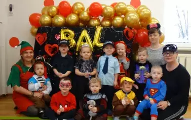 Sala zabaw-Opiekunowie i grupa dzieci w kolorowych balowych strojach, jedni stoją drudzy siedzą na dywanie. W tle kolorowe balony i załoty napis BAL.