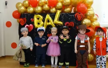 Sala zabaw-grupa dzieci w kolorowych balowych strojach stoi na dywanie. W tle kolorowe balony i załoty napis BAL.