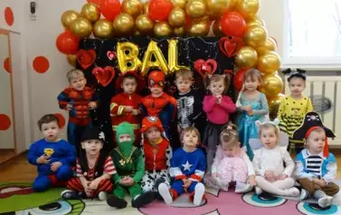 Sala zabaw-grupa dzieci w kolorowych balowych strojach, jedni stoją drudzy siedzą na dywanie. W tle kolorowe balony i załoty napis BAL.