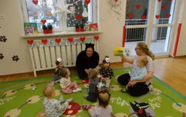 Sala zabaw-Opiekun z grupą dzieci siedzi na dywanie. Na kolanach trzyma dziecko a w ręce książeczkę. Obok nich znajduje się drugi Opiekun w przebraniu kota.