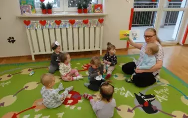 Sala zabaw-Opiekun z grupką dzieci siedzi na dywanie. Na kolanach trzyma dziecko a w ręce książeczkę.