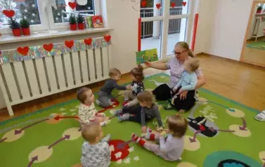 Sala zabaw-Opiekun z grupką dzieci siedzi na dywanie. Na kolanach trzyma dziecko a w ręce książeczkę.