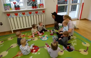 Sala zabaw-Opiekun z grupą dzieci siedzi na dywanie. Obok nich znajduje się drugi Opiekun w przebraniu kota.