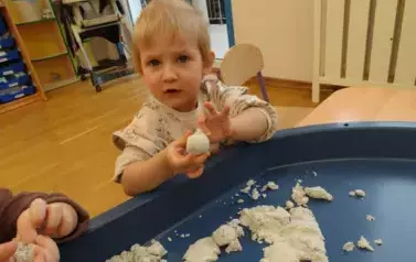 Chłopiec siedzi przy stoliku z podciągniętymi do łokcia rękawkami. Tuż przed nim leży granatowa taca z piaskiem. Dziecko ma w ręku kulkę z piasku. W tle sala zabaw.