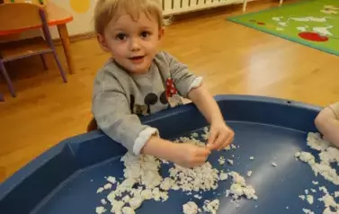 Na pierwszym planie dziecko w szarej bluzie z podciągniętymi do łokcia rękawkami. Tuż przed nim granatowa taca z piaskiem. Dziecko dotyka rękoma piasek. W tle sala zabaw.