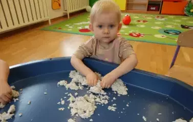 Na pierwszym planie dziecko w kolorowym ubranku z podciągniętymi do łokcia rękawkami. Tuż przed nim granatowa taca z piaskiem. Dziecko dotyka rękoma piasek. W tle sala zabaw.