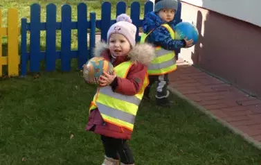 Dziewczynka z chłopcem stoją na zielonej trawie. W ręce trzymają kolorowe piłki.