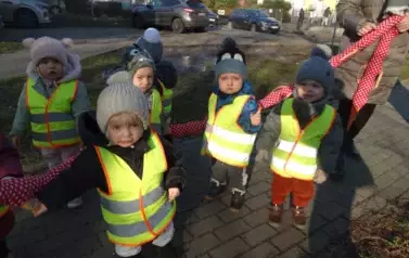 Grupa dzieci stoi przed budynkiem przy wężu spacerowym. Na swoją odzież mają założone odblaskowe kamizelki.