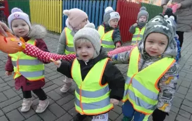 Grupa dzieci stoi przed budynkiem przy wężu spacerowym. Na swoją odzież mają założone odblaskowe kamizelki.
