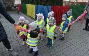 Grupa dzieci stoi przed budynkiem przy wężu spacerowym. Na swoją odzież mają założone odblaskowe kamizelki.