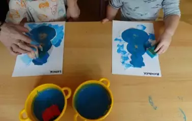 Sala zabaw-dzieci siedzą przy stoliku, na kt&oacute;rym leżą kartki papieru. W ręce trzymają niebieskie gąbki, kt&oacute;rymi dotykają kartek.
