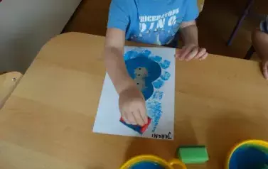Sala zabaw-dziecko siedzi przy stoliku, na kt&oacute;rym leży kartka papieru. W ręce trzyma gąbkę, kt&oacute;rą dotyka kartki.