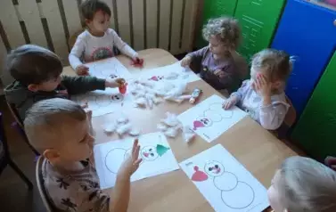 Sala zabaw-dzieci siedzą przy stoliku, na kt&oacute;rym leżą kartki papieru z konturami bałwank&oacute;w i biała wata.