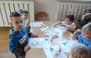 Sala zabaw-dzieci siedzą przy stoliku, na kt&oacute;rym leżą kartki papieru z konturami bałwank&oacute;w i biała wata.