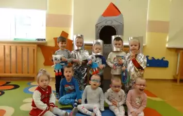 Sala zabaw-na pierwszym planie grupka dzieci siedzi na dywanie. Tuż za nimi stoi szaro-czerwony statek kosmiczny a przy nim chłopcy w przebraniu kosmonat&oacute;w.