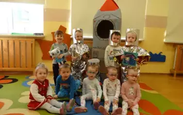 Sala zabaw-na pierwszym planie grupka dzieci siedzi na dywanie. Tuż za nimi stoi szaro-czerwony statek kosmiczny a przy nim chłopcy w przebraniu kosmonat&oacute;w.