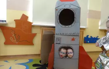 Sala zabaw-na dywanie stoi szaro-czerwony statek kosmiczny. Ma dwa okrągłe okienka. W jednym z nich widać dwie twarze dzieci.