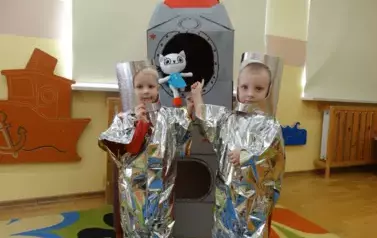 Sala zabaw-dw&oacute;ch chłopc&oacute;w przebranych za kosmonaut&oacute;w stoi na dywanie. Tuż za nimi znajduje się statek kosmiczny.
